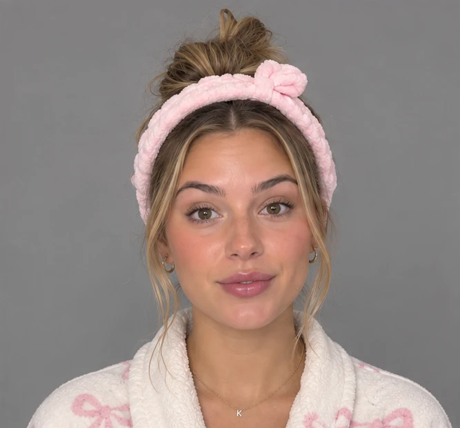 Chloe's front portrait on a plain grey background: hair in a bun under a pink knotted headband, small gold hoops, a fine necklace with a K pendant and a cream robe with pink bows.